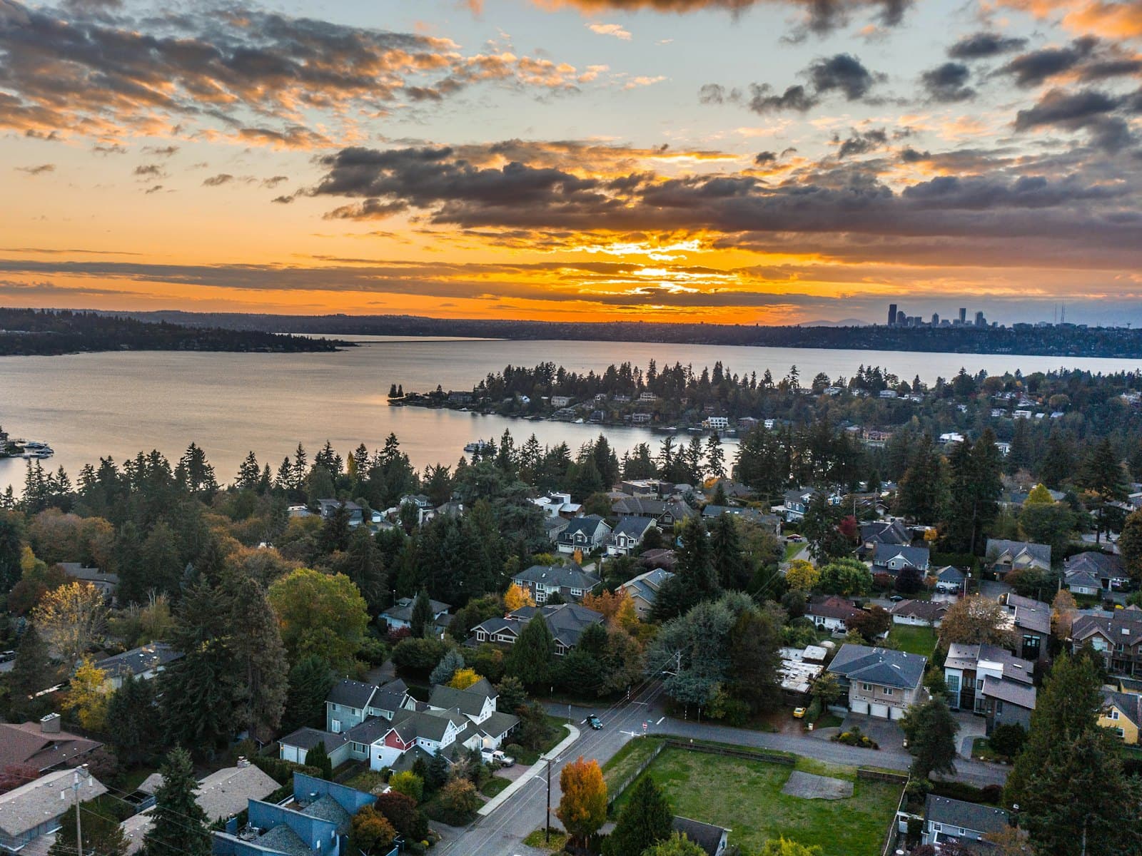 Homes among trees above a lake at sunset, with a city skyline on the far shore