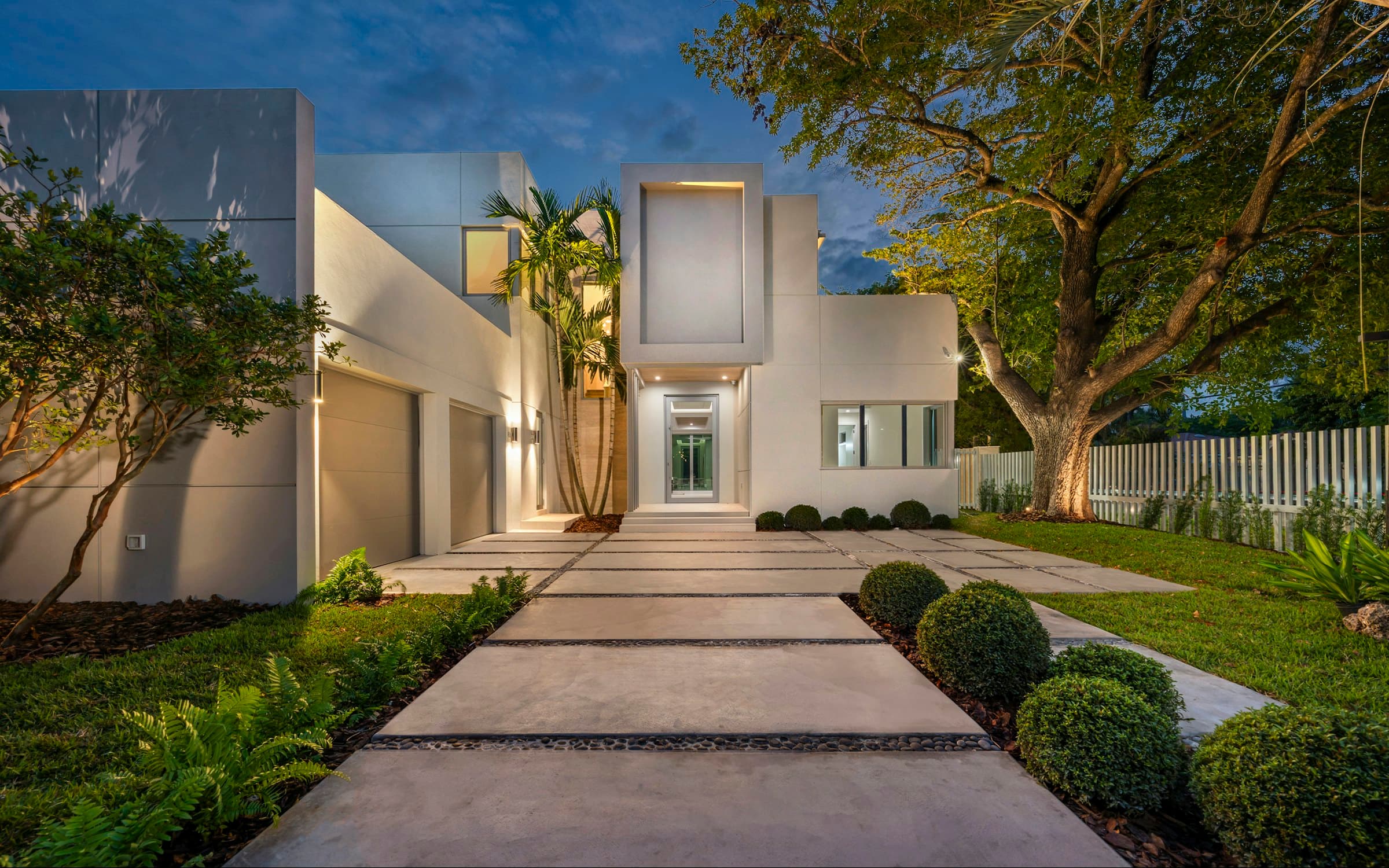 A modern white home lit up at dusk, with a stone path leading to the front door